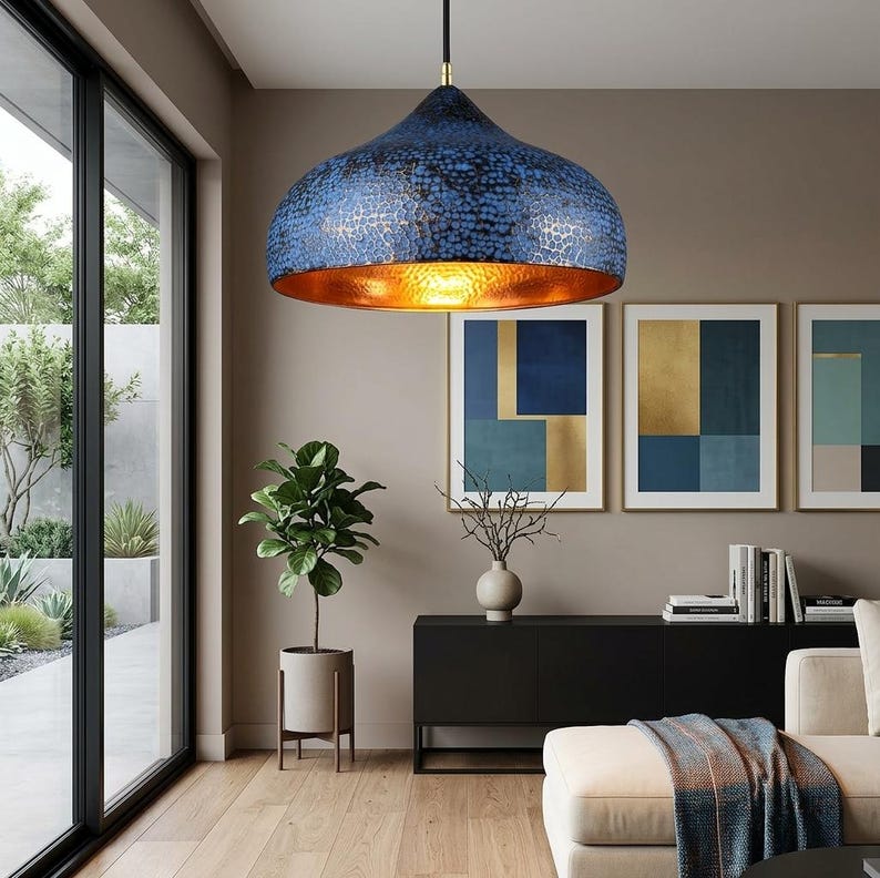 May include: A blue and gold pendant light fixture with a textured surface hangs from the ceiling. The interior of the light is gold, and the fixture is suspended above a living room with artwork and a sofa.
