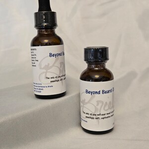 Op de afbeelding: Twee amberkleurige glazen flessen met zwarte druppelaars en witte etiketten. De etiketten hebben de tekst "Beyond Beard Oil" en "De enige olie die je ooit nodig zult hebben om een ​​verbluffend zachte en boeiende baard te laten groeien."