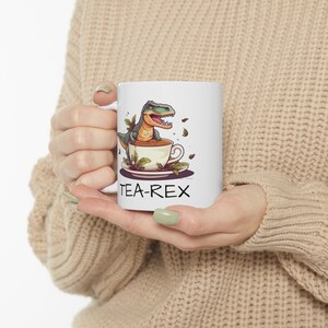 May include: White ceramic mug with a green dinosaur drinking tea from a teacup. The text "TEA-REX" is printed on the mug.