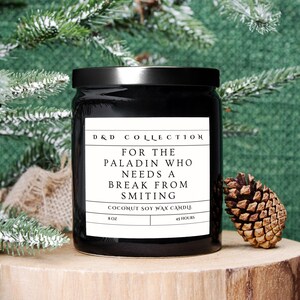 May include: Black candle jar with a white label that reads "D&D COLLECTION FOR THE PALADIN WHO NEEDS A BREAK FROM SMITING COCONUT SOY WAX CANDLE 8OZ 45 HOURS".