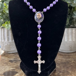 May include: A rosary necklace featuring smooth, round, light purple beads linked by silver-toned metal. The center piece is an oval pendant containing a small photograph of a person wearing glasses and a plaid shirt, ending in a silver-toned crucifix.