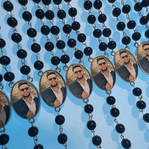 May include: Multiple rosaries featuring glossy black beads connected by silver-toned metal links. Each rosary includes an oval-shaped pendant displaying a photographic portrait of a person wearing sunglasses, a white collared shirt, and a dark blazer.