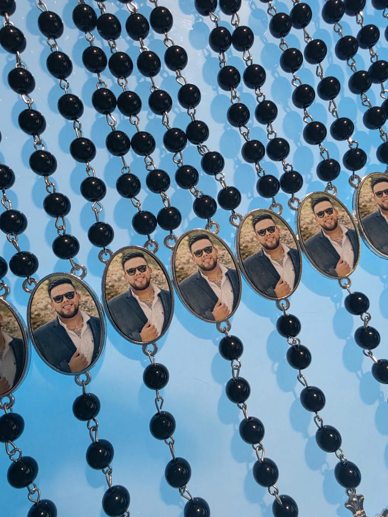 May include: Multiple rosaries featuring glossy black beads connected by silver-toned metal links. Each rosary includes an oval-shaped pendant displaying a photographic portrait of a person wearing sunglasses, a white collared shirt, and a dark blazer.