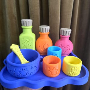 May include: A set of colorful silicone play items on a purple tray, including a lime green bottle with a leaf, an orange bottle with a moon, a pink bottle with a heart, a purple cauldron with a yellow spoon, and three small cups with star and flower icons.