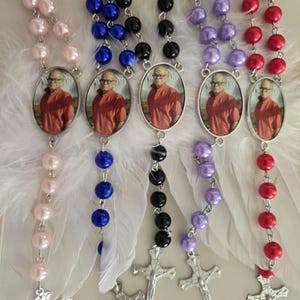 May include: Five rosaries featuring colored pearl beads in light pink, blue, black, purple, and red. Each rosary includes a silver-toned metal crucifix and an oval pendant containing a photograph of a person wearing an orange shirt. White feathers are attached to the center of each rosary.