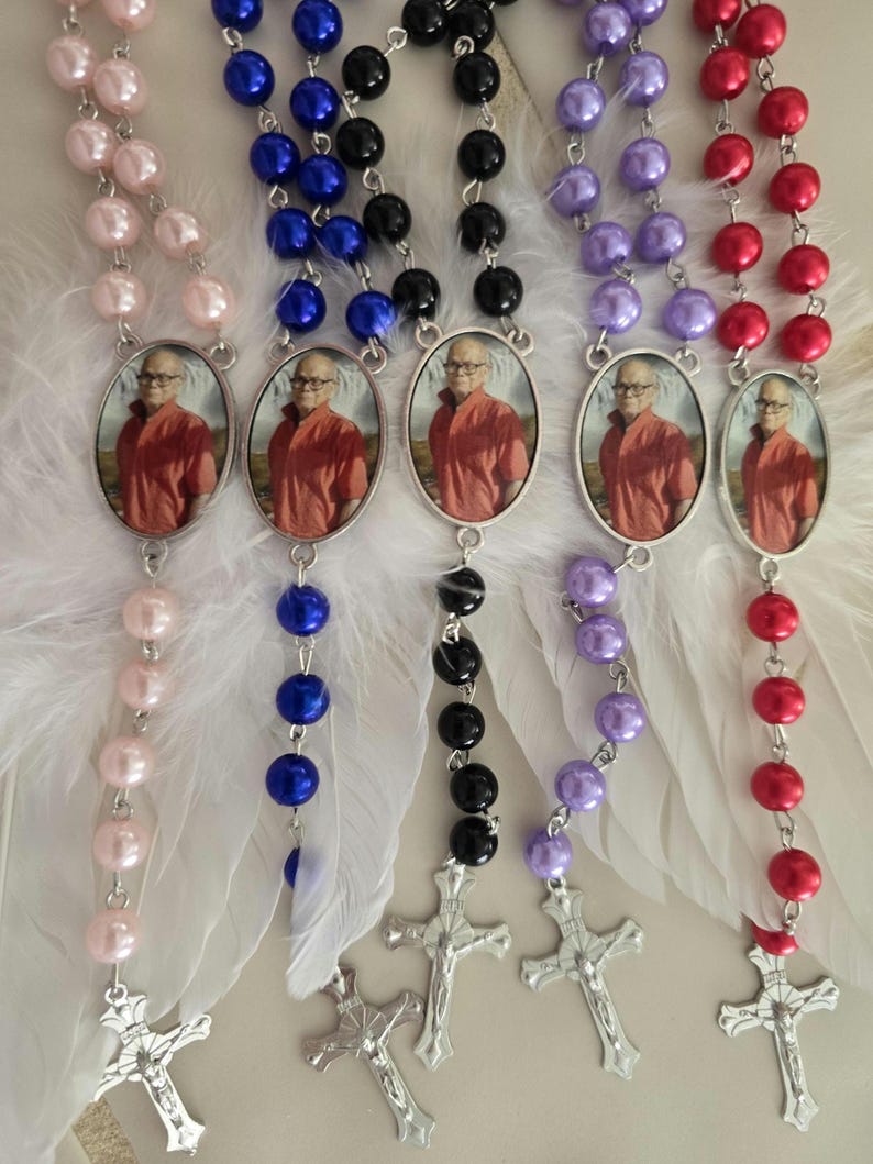 May include: Five rosaries featuring colored pearl beads in light pink, blue, black, purple, and red. Each rosary includes a silver-toned metal crucifix and an oval pendant containing a photograph of a person wearing an orange shirt. White feathers are attached to the center of each rosary.