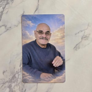 May include: A rectangular memorial card featuring a portrait of a bald, mustachioed individual wearing glasses and a dark blue long-sleeved shirt. The person is positioned against a background of a bright blue sky filled with soft white clouds.
