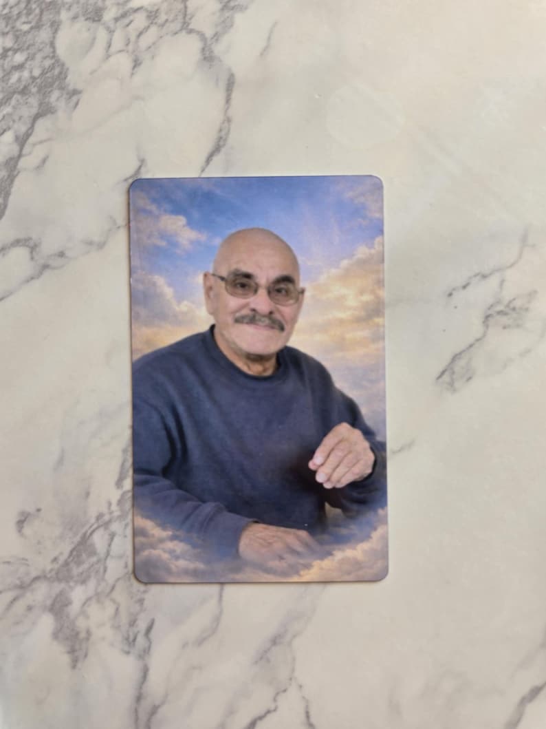 May include: A rectangular memorial card featuring a portrait of a bald, mustachioed individual wearing glasses and a dark blue long-sleeved shirt. The person is positioned against a background of a bright blue sky filled with soft white clouds.