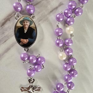 May include: A rosary featuring lustrous lavender beads, interspersed with two white beads, connected by silver-toned metal links. An oval pendant displays a photograph of a person wearing a blue hat and dark vest, and a silver crucifix hangs at the end.