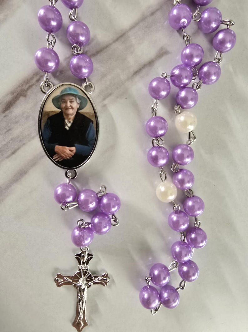 May include: A rosary featuring lustrous lavender beads, interspersed with two white beads, connected by silver-toned metal links. An oval pendant displays a photograph of a person wearing a blue hat and dark vest, and a silver crucifix hangs at the end.
