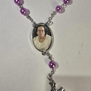 May include: A rosary necklace with purple beads and a silver cross pendant. The necklace features an oval pendant with a photograph of a woman. The beads are connected by a silver chain.