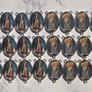 May include: A collection of twenty-seven oval metal charms arranged in three rows. The charms feature two alternating religious images: one depicting a crowned figure in robes and another showing a group of three figures with halos in blue tones.