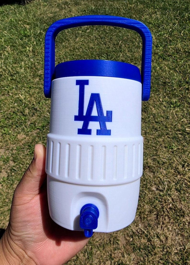 May include: A white and blue plastic beverage cooler featuring a blue handle, a blue spigot at the base, and the blue LA logo printed on the front. The cylindrical container has a ribbed midsection and a solid blue lid, designed for holding liquids.