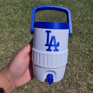 May include: A white plastic beverage cooler with a blue handle, blue lid, and a blue spigot at the base. The front of the cooler features the blue Los Angeles Dodgers LA logo. The cylindrical container has a ribbed texture around the middle section.