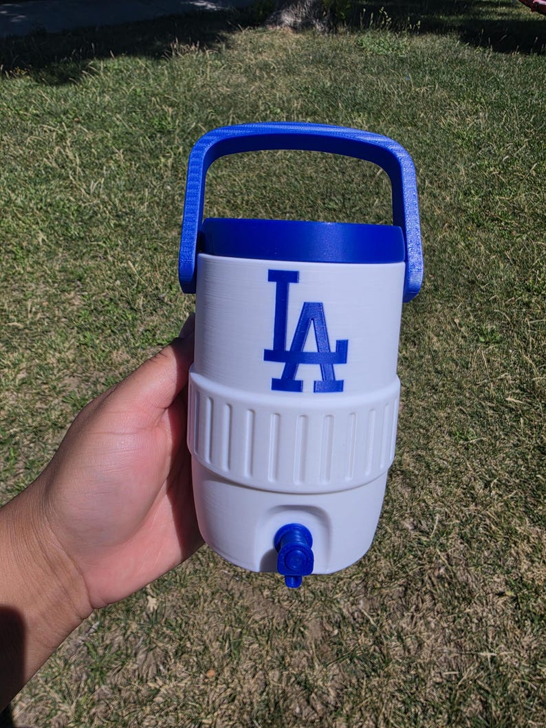 May include: A white plastic beverage cooler with a blue handle, blue lid, and a blue spigot at the base. The front of the cooler features the blue Los Angeles Dodgers LA logo. The cylindrical container has a ribbed texture around the middle section.