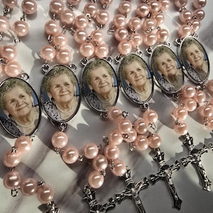 May include: A collection of rosaries featuring light pink faux pearl beads, silver-toned metal crucifixes, and oval-shaped pendants containing a black and white photographic portrait of an elderly person wearing a necklace.