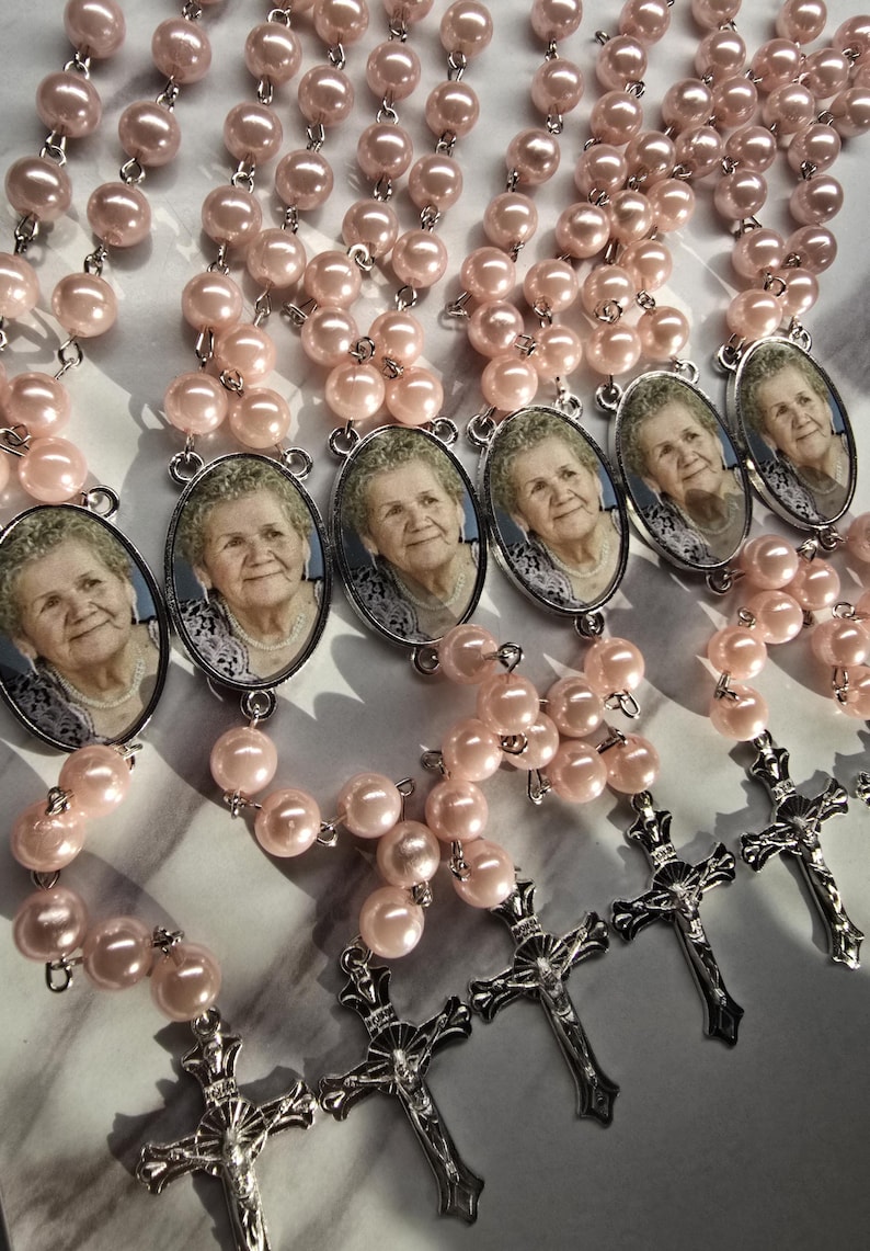 May include: A collection of rosaries featuring light pink faux pearl beads, silver-toned metal crucifixes, and oval-shaped pendants containing a black and white photographic portrait of an elderly person wearing a necklace.
