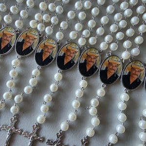 May include: Multiple rosaries featuring white faux pearl beads connected by silver-toned metal links. Each rosary includes an oval-shaped pendant displaying a photograph of a person wearing a cowboy hat and a black shirt, finished with a silver crucifix.