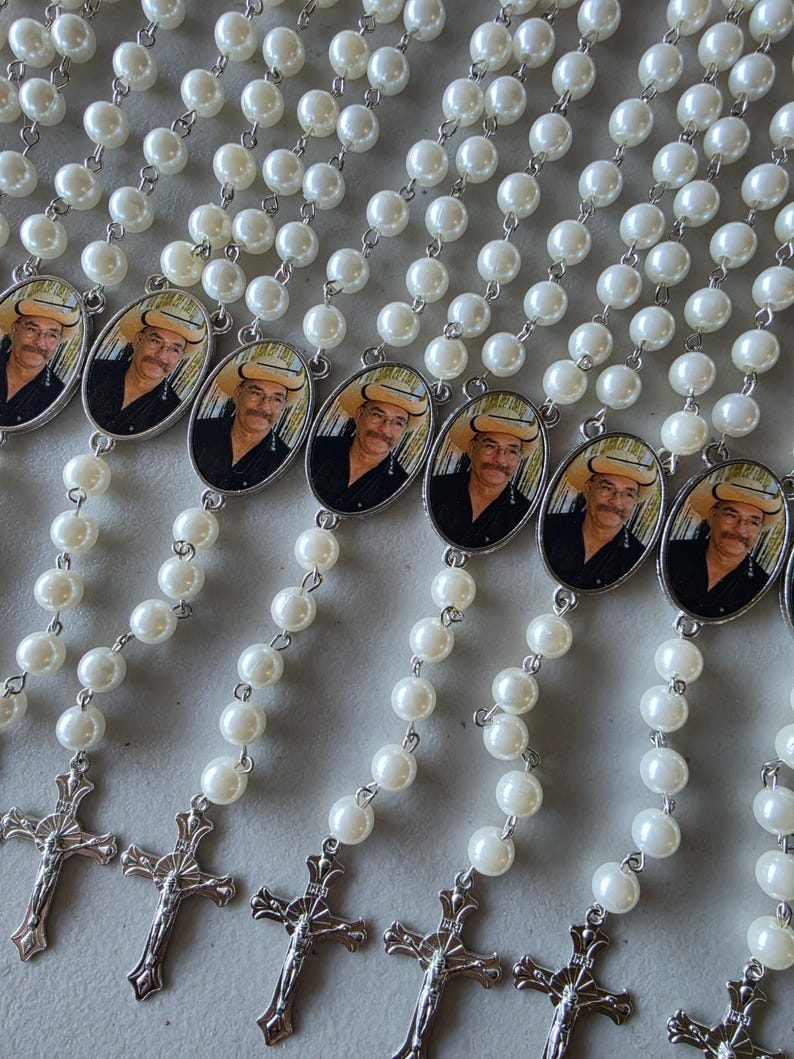 May include: Multiple rosaries featuring white faux pearl beads connected by silver-toned metal links. Each rosary includes an oval-shaped pendant displaying a photograph of a person wearing a cowboy hat and a black shirt, finished with a silver crucifix.