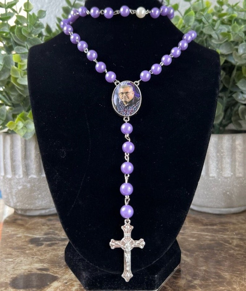 May include: A rosary necklace featuring smooth, round, light purple beads linked by silver-toned metal. The center piece is an oval pendant containing a small photograph of a person wearing glasses and a plaid shirt, ending in a silver-toned crucifix.