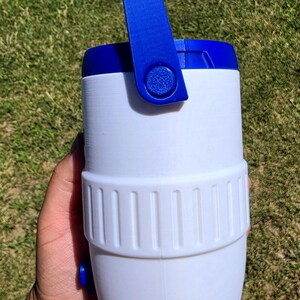 May include: A white cylindrical container with a blue lid and a blue pivoting handle. The body features a textured middle band with vertical ridges and a small blue circular protrusion on the lower side, constructed from a smooth plastic material.