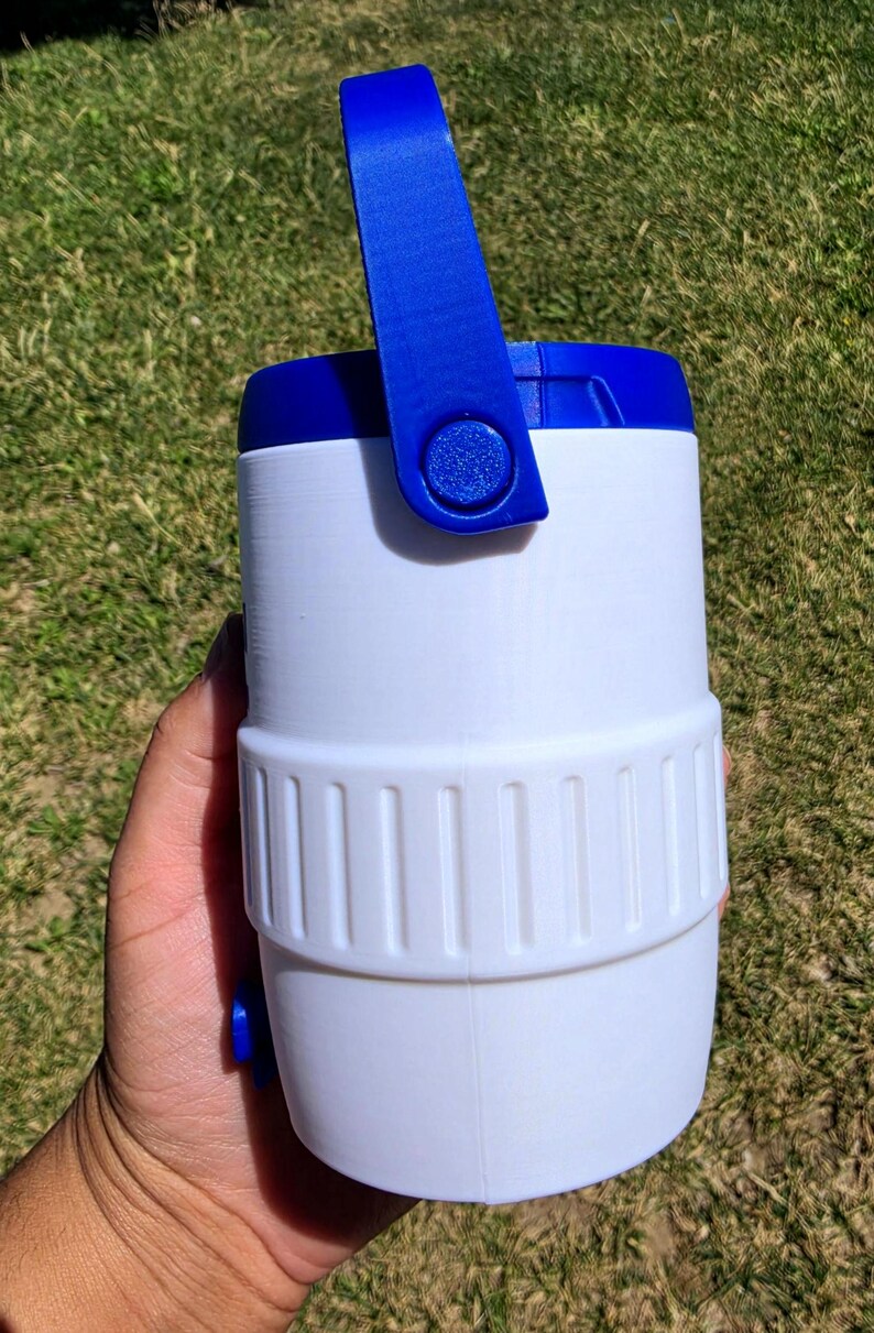 May include: A white cylindrical container with a blue lid and a blue pivoting handle. The body features a textured middle band with vertical ridges and a small blue circular protrusion on the lower side, constructed from a smooth plastic material.