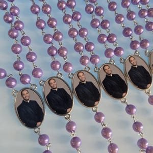 May include: Multiple strands of rosary beads featuring round, glossy, light purple faux pearls connected by silver-toned metal links. Each strand includes an oval-shaped metal pendant displaying a photographic portrait of a smiling person in a black suit.