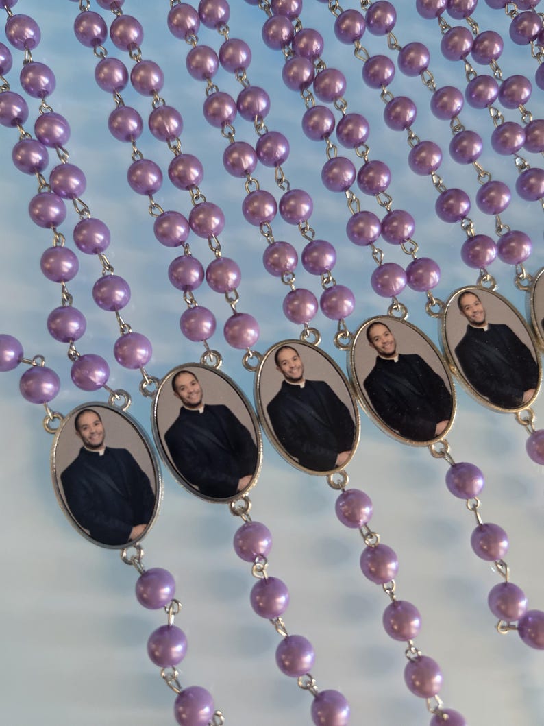 May include: Multiple strands of rosary beads featuring round, glossy, light purple faux pearls connected by silver-toned metal links. Each strand includes an oval-shaped metal pendant displaying a photographic portrait of a smiling person in a black suit.