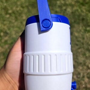 May include: A white and blue plastic beverage cooler featuring a cylindrical body with a textured middle band, a blue top lid, a blue swing handle attached to the sides, and a small blue spigot located near the bottom of the container.
