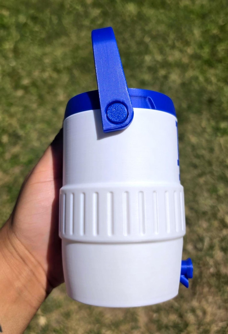 May include: A white and blue plastic beverage cooler featuring a cylindrical body with a textured middle band, a blue top lid, a blue swing handle attached to the sides, and a small blue spigot located near the bottom of the container.