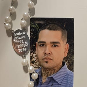 May include: A memorial rosary featuring white faux pearl beads and a silver-toned oval pendant engraved with the text Ruben Mares Jr 1993-2025, positioned next to a rectangular photograph of a person with short dark hair and a neck tattoo.