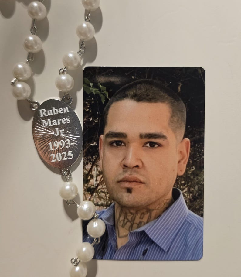 May include: A memorial rosary featuring white faux pearl beads and a silver-toned oval pendant engraved with the text Ruben Mares Jr 1993-2025, positioned next to a rectangular photograph of a person with short dark hair and a neck tattoo.