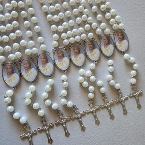 May include: A collection of eight rosaries featuring white faux pearl beads linked by silver-toned metal chains. Each rosary includes an oval-shaped pendant containing a photograph of an older person wearing a yellow shirt, finished with a silver crucifix.