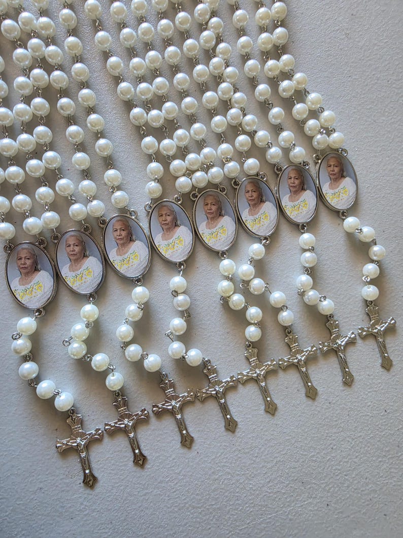 May include: A collection of eight rosaries featuring white faux pearl beads linked by silver-toned metal chains. Each rosary includes an oval-shaped pendant containing a photograph of an older person wearing a yellow shirt, finished with a silver crucifix.