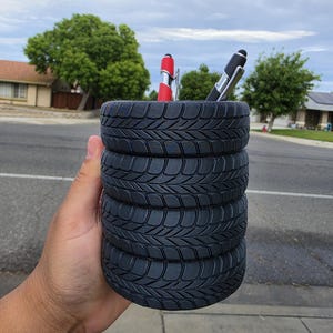 May include: A cylindrical desk organizer shaped like a stack of four black rubber tires with deep tread patterns. The hollow center holds two pens, one with a red barrel and one with a black barrel, both featuring silver clips and stylus tips.