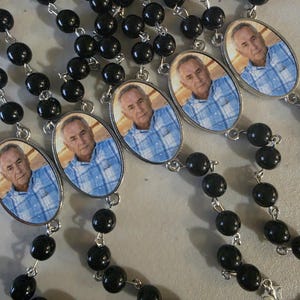 May include: A collection of rosaries featuring black beads connected by silver-toned metal links. Each rosary includes an oval-shaped pendant displaying a photographic portrait of an older man wearing a blue and white plaid button-down shirt.