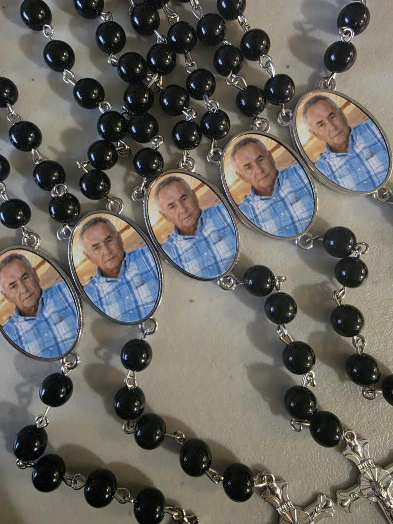May include: A collection of rosaries featuring black beads connected by silver-toned metal links. Each rosary includes an oval-shaped pendant displaying a photographic portrait of an older man wearing a blue and white plaid button-down shirt.