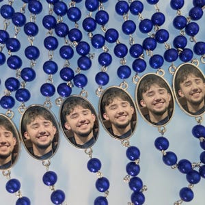 May include: A collection of rosaries featuring vibrant royal blue spherical beads connected by silver-toned metal links. Each rosary includes an oval-shaped pendant displaying a photographic portrait of a smiling person with dark hair.