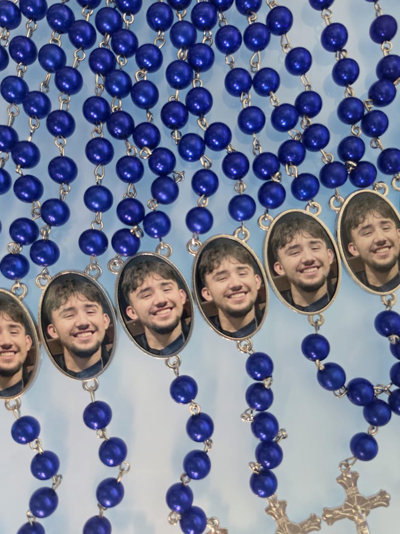 May include: A collection of rosaries featuring vibrant royal blue spherical beads connected by silver-toned metal links. Each rosary includes an oval-shaped pendant displaying a photographic portrait of a smiling person with dark hair.
