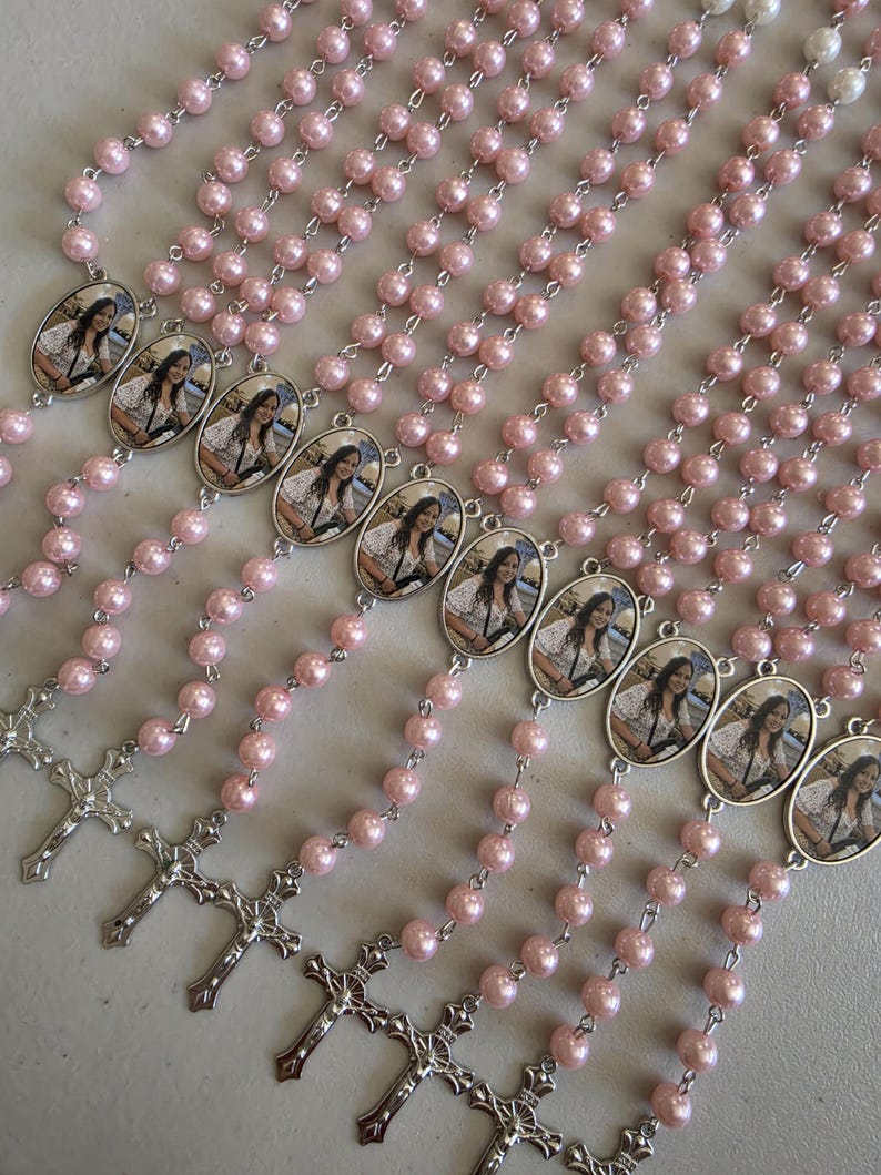 May include: A collection of rosaries featuring light pink pearl beads and silver-toned metal chains. Each rosary includes an oval-shaped pendant containing a photographic portrait of a person and a silver-toned crucifix at the bottom end.