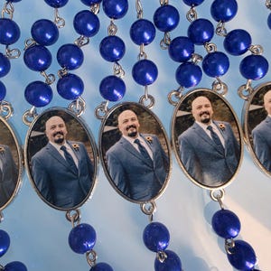 May include: A series of rosaries featuring glossy, deep blue spherical beads linked by silver-toned metal wire. Each rosary includes an oval-shaped pendant containing a photographic portrait of a person with a shaved head and beard wearing a suit.