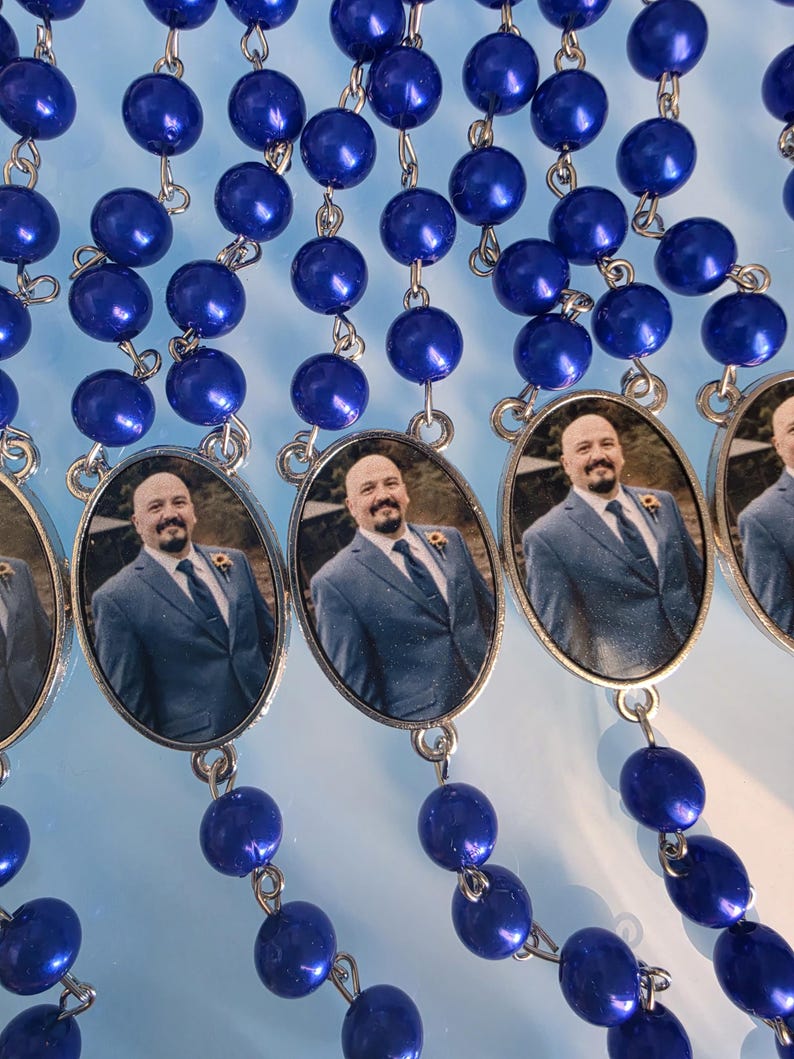 May include: A series of rosaries featuring glossy, deep blue spherical beads linked by silver-toned metal wire. Each rosary includes an oval-shaped pendant containing a photographic portrait of a person with a shaved head and beard wearing a suit.