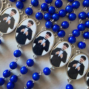 May include: A collection of rosaries featuring vibrant royal blue beads connected by silver-toned metal links. Each rosary includes an oval-shaped pendant containing a photographic portrait of a person wearing a black tuxedo with a bow tie.