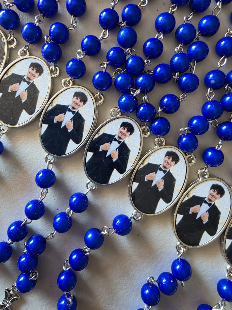 May include: A collection of rosaries featuring vibrant royal blue beads connected by silver-toned metal links. Each rosary includes an oval-shaped pendant containing a photographic portrait of a person wearing a black tuxedo with a bow tie.