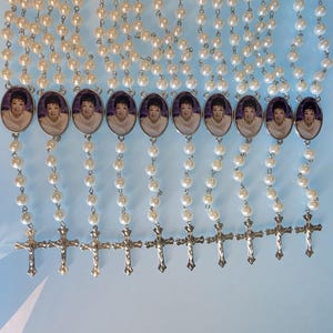 May include: A collection of ten rosaries featuring white faux pearl beads linked by silver-toned metal chains. Each rosary includes an oval-shaped pendant displaying a portrait of a person and a silver-toned metal crucifix at the bottom end.