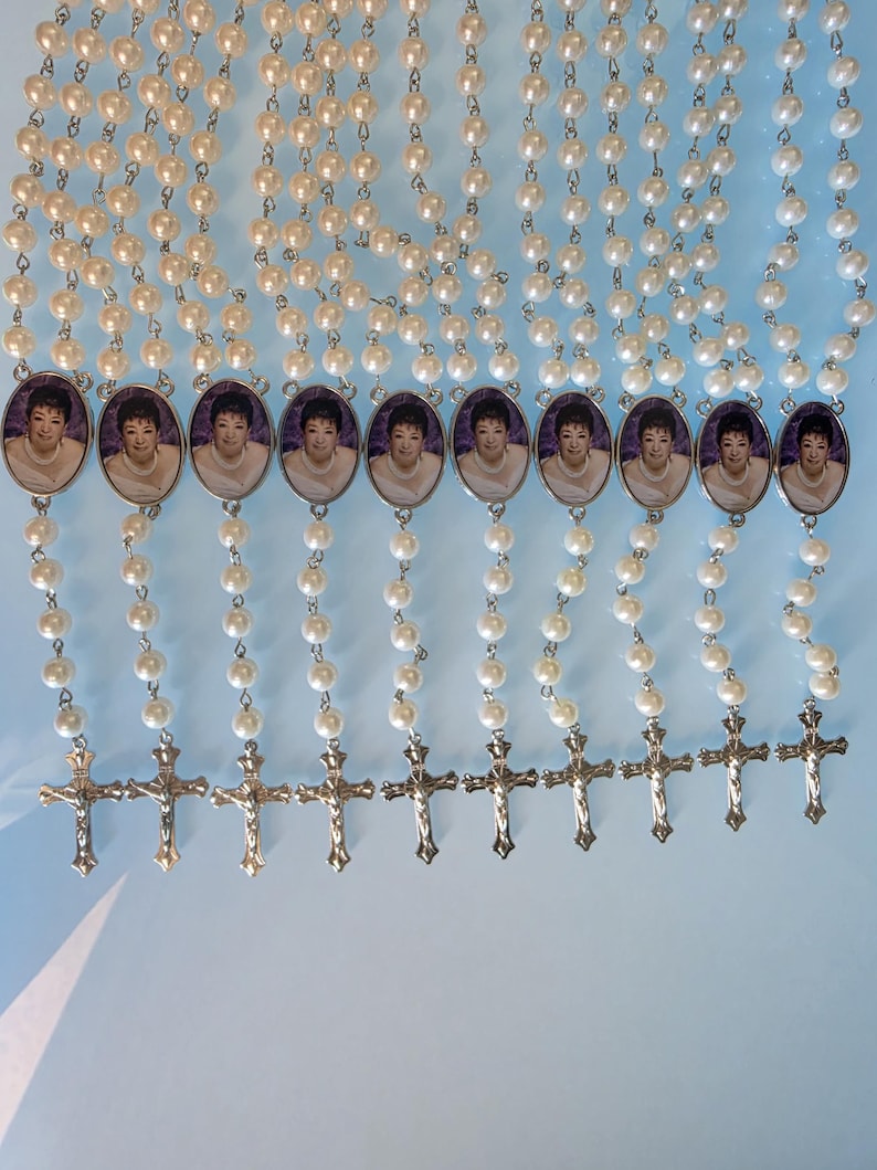 May include: A collection of ten rosaries featuring white faux pearl beads linked by silver-toned metal chains. Each rosary includes an oval-shaped pendant displaying a portrait of a person and a silver-toned metal crucifix at the bottom end.