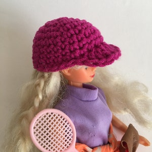 Könnte beinhalten: Eine Puppe mit blonden Haaren, die einen rosa gehäkelten Hut mit Krempe und ein lila Shirt trägt. Die Puppe hält einen rosa Tennisschläger.