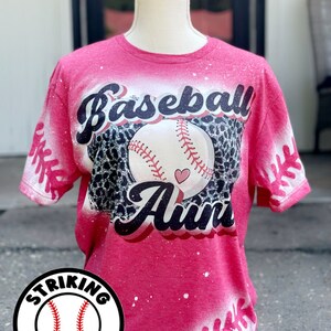 May include: Pink t-shirt with a bleach-splattered design featuring a baseball with a heart and the text "Baseball Aun." The shirt also has a white baseball stitch design on the sleeves and bottom. The shirt is on a white mannequin. A white circle with a black baseball and the text "Striking Impressions" is in the bottom left corner.