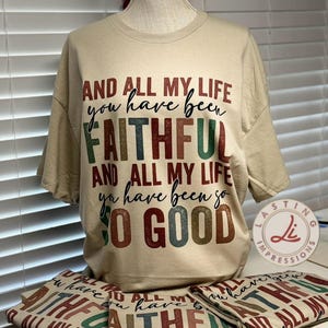 Könnte beinhalten: Beiges Kurzarm-T-Shirt mit dem Text "AND ALL MY LIFE you have been FAITHFUL AND ALL MY LIFE you have been so GOOD" in verschiedenen Farben. Das Shirt wird auf einer Schaufensterpuppe und gefaltet auf einem Tisch präsentiert.
