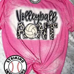 May include: Pink bleached t-shirt with a white volleyball and the text "Volleyball Aunt" in black with a leopard print background. The shirt has a white splatter design and a logo for "Striking Impressions" in the top left and bottom right corners.
